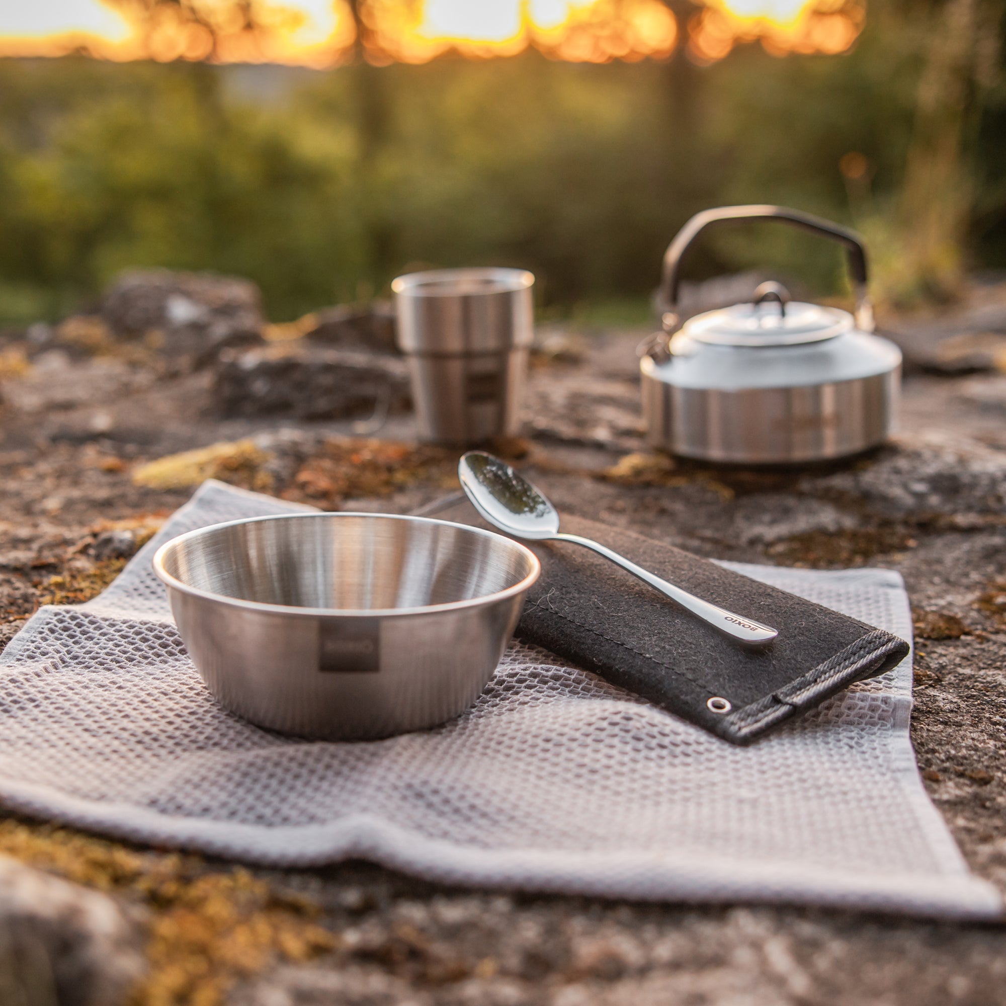Waldszenerie mit einer Edelstahlschüssel, einem Löffel, Bestecktasche, im Hintergrund ist ein Edelstahlbecher und ein Edelstahlwasserkessel zu sehen