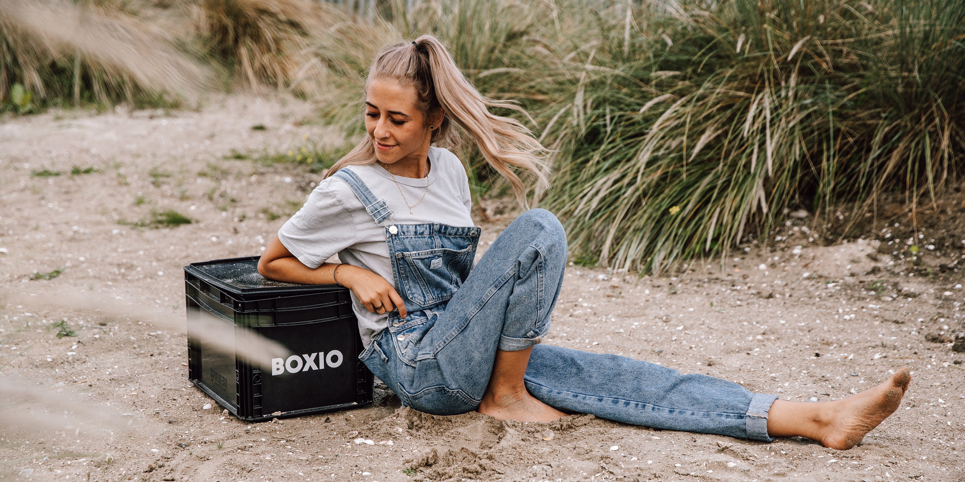 Junge Frau in Latzhose sitzt entspannt im Sand neben einer schwarzen Kiste mit dem Aufdruck "BOXIO".