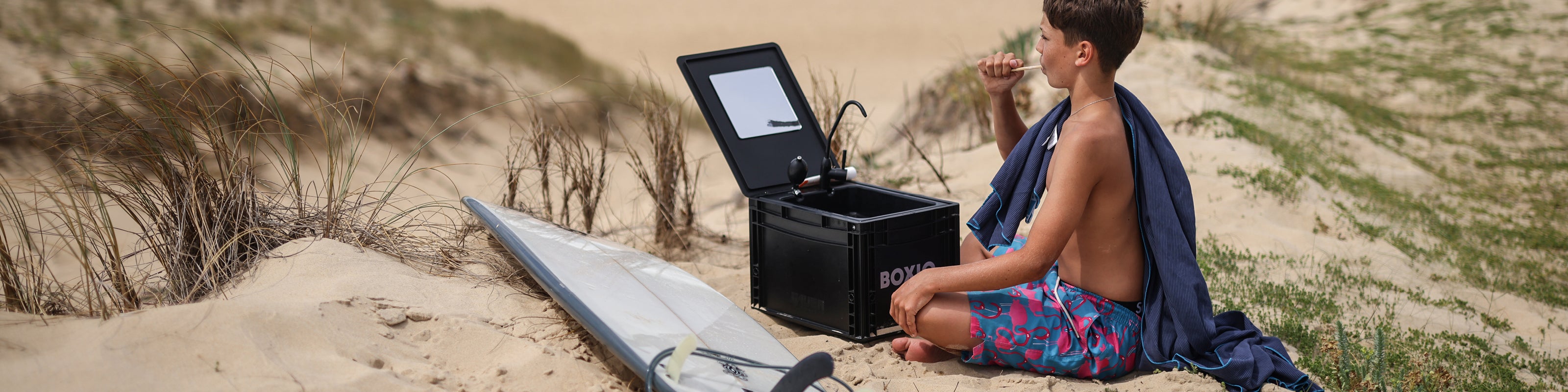 Junge sitzt im Sand am Strand, neben einem offenen Kühlbox, betont lässiger Sommerlook mit Surfbrett im Hintergrund.