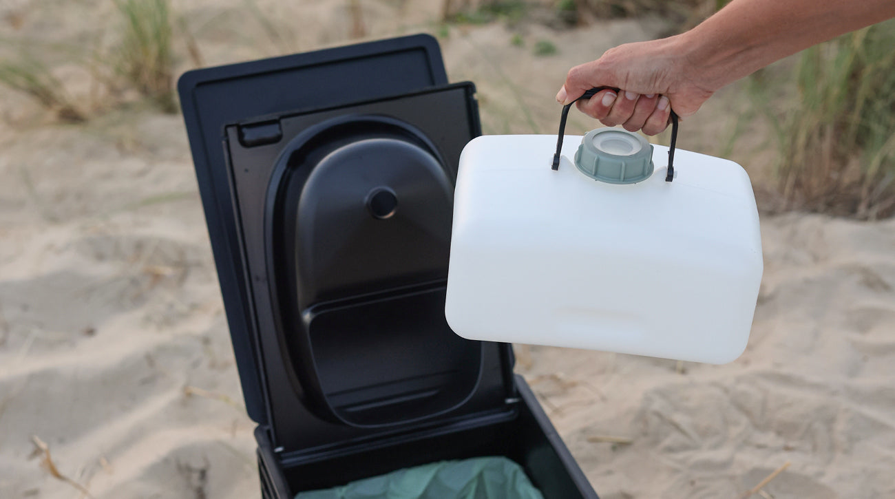 Eine Hand hält einen weißen Kanister über einer geöffneten tragbaren Toilette auf sandigem Untergrund.