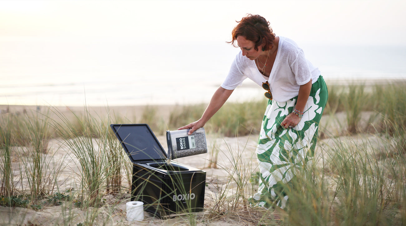Frau in Strandkleidung gießt einen Shake in eine schwarze Boxio-Box am Strand, umgeben von Strandgras.