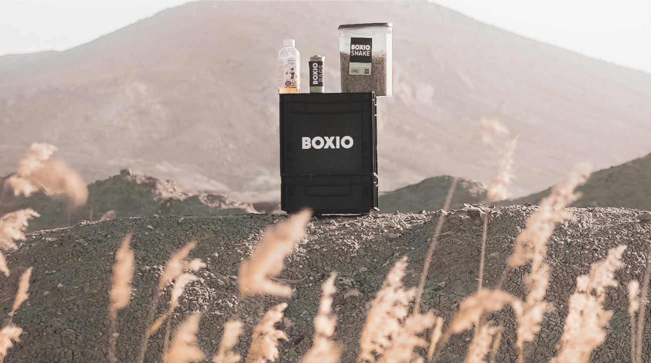 Schwarze Box mit der Aufschrift "BOXIO", daneben Nahrungsergänzungsmittel auf einem hügeligen Untergrund.