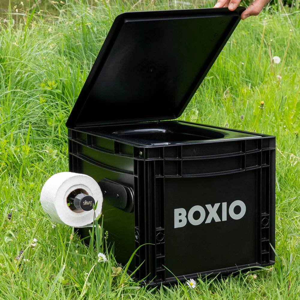 Schwarze Outdoor-Toilette mit geöffnetem Deckel und integriertem Toilettenpapierhalter auf einer Wiese.